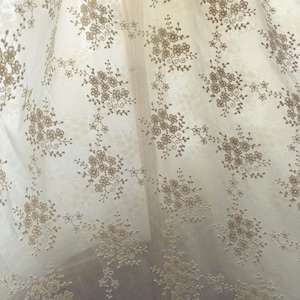 Pu&ograve; includere: Tessuto trasparente bianco sporco con un motivo ripetuto di gruppi floreali ricamati in marrone chiaro. Il design delicato crea un'estetica morbida e ariosa, adatta per tende o tessuti decorativi.