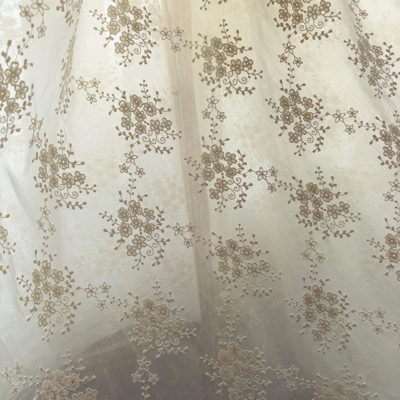 Pu&ograve; includere: Tessuto trasparente bianco sporco con un motivo ripetuto di gruppi floreali ricamati in marrone chiaro. Il design delicato crea un'estetica morbida e ariosa, adatta per tende o tessuti decorativi.