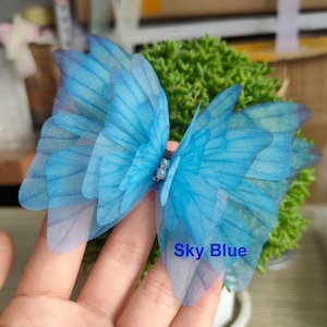 May include: A sky blue butterfly hair clip with layered, translucent wings. The wings have delicate veining details and are attached to a small, beaded center. The text "Sky Blue" is visible on the image.