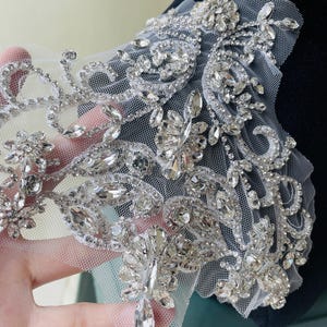 Puede incluir: Primer plano de una delicada aplicación de encaje. La aplicación presenta un diseño floral complejo con brillantes pedrería y cristales transparentes de varios tamaños, sobre una tela de malla blanca transparente. El diseño es intrincado y detallado, con un borde festoneado.