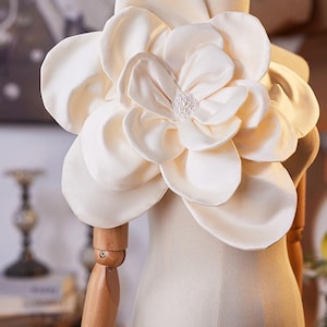 Puede incluir: Una flor de tela grande de color crema con pétalos redondeados en capas y un centro decorado con un grupo de pequeñas perlas artificiales blancas. El accesorio se exhibe sobre un torso de maniquí de madera, mostrando su diseño estructurado.