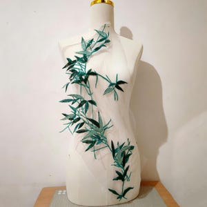 May include: A sheer white fabric appliqué featuring an embroidered design of dark green bamboo leaves and stems. The botanical pattern is arranged in a vertical, flowing composition, with detailed stitching and texture on the leaves.