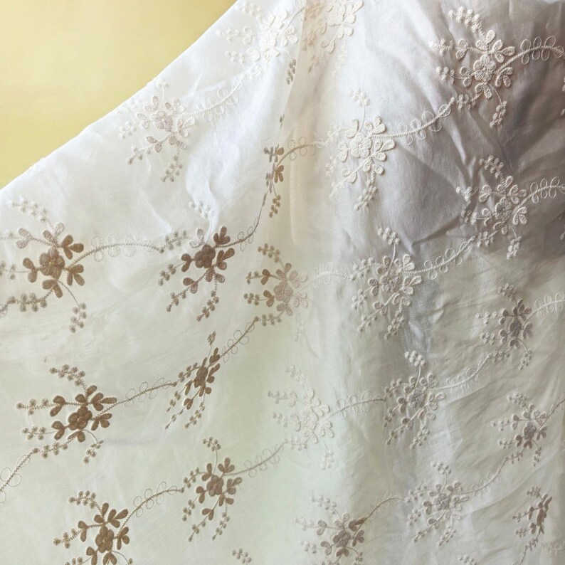 May include: Close-up of white fabric with embroidered floral patterns in a light brown color. The fabric is slightly translucent, with the floral designs arranged in a repeating pattern across the surface. The background is a soft yellow.