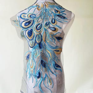 May include: An embroidered peacock feather design on sheer fabric. The design uses blue, gold, and navy sequins and thread, creating a shimmering effect. The embroidery is detailed and intricate, with a focus on the feather's structure.