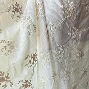 May include: Close-up of white fabric with intricate floral embroidery. The embroidery features delicate flowers and leaves in a contrasting light brown color. The fabric is slightly wrinkled, adding texture to the design. The overall effect is elegant and detailed.