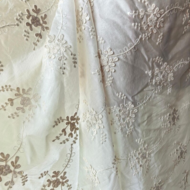 May include: Close-up of white fabric with intricate floral embroidery. The embroidery features delicate flowers and leaves in a contrasting light brown color. The fabric is slightly wrinkled, adding texture to the design. The overall effect is elegant and detailed.