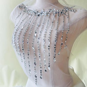 May include: A sheer white fabric adorned with silver-colored rhinestones and beads. The embellishments are arranged in a decorative pattern around the neckline and cascade down in vertical strands, creating a shimmering effect. The design is displayed on a mannequin.