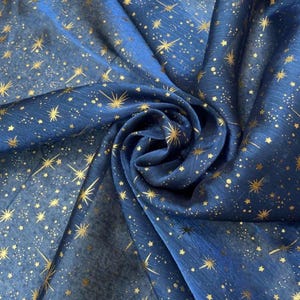 Puede incluir: Una tela azul oscuro adornada con patrones dorados de estrellas y puntos, creando un tema celestial. La tela tiene una calidad brillante, con un efecto degradado de azul claro a oscuro, y está arremolinada.