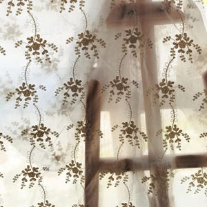 May include: Sheer white curtain with a repeating pattern of embroidered brown floral sprigs. The curtain is hanging in front of a window, allowing light to filter through. The floral design adds a delicate, natural touch.