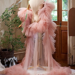 May include: A dusty pink tulle robe featuring voluminous, layered ruffled sleeves and a long, flowing train. The garment is displayed on a mannequin, showcasing the sheer, pleated fabric construction and the extensive, gathered tulle detailing.