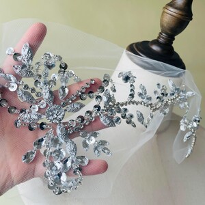 以下が含まれることがあります： 銀色のビーズアップリケ、フローラルデザイン。透明でファセットカットされたクリスタル、スパンコール、ビーズが、透明な白いメッシュの裏地に配置されています。デザインには葉と花の形が含まれており、きらびやかな装飾要素を作り出しています。