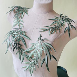 Puede incluir: Aplicaciones de hojas bordadas en tonos verdes y blancos, dispuestas sobre un maniquí. Las aplicaciones presentan costuras detalladas, que se asemejan a un delicado follaje. Ideales para embellecer prendas o proyectos de artesanía, estas aplicaciones añaden un toque de elegancia inspirada en la naturaleza.