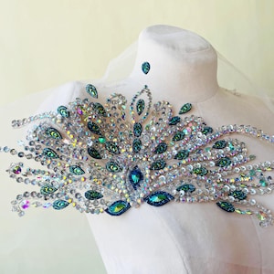 Puede incluir: Un diseño decorativo de pluma de pavo real iridiscente, adornado con pedrería transparente y verde azulado. El diseño está sobre una base de tela blanca, creando un efecto brillante y llamativo. La pieza probablemente está destinada a adornar ropa o accesorios.