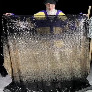 May include: A large piece of fabric with a gradient effect, transitioning from black at the bottom to gold in the middle and sparkling at the top. The fabric is held up, showcasing its shimmering sequins.