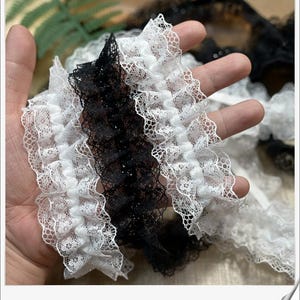 May include: Close-up of three ruffled lace trims in white and black. The lace has a delicate, openwork design with a scalloped edge. The trims are held in a hand, showcasing the texture and detail of the fabric. Ideal for sewing and craft projects.