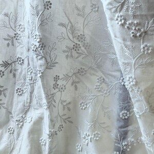 May include: White fabric with intricate floral embroidery. The design features delicate embroidered flowers and leaves, creating a textured and detailed pattern. The fabric appears soft and flowing, with visible folds.