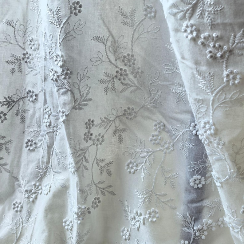 May include: White fabric with intricate floral embroidery. The design features delicate embroidered flowers and leaves, creating a textured and detailed pattern. The fabric appears soft and flowing, with visible folds.