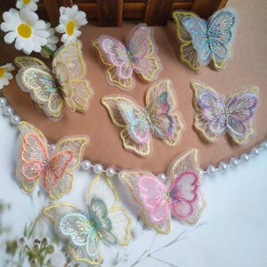 Può includere: Una collezione di applicazioni a farfalla ricamate colorate. Ogni farfalla presenta delicate ali trasparenti con cuciture intricate in tonalità di rosa, giallo, blu e verde, delineate con filo dorato. Questi abbellimenti decorativi sono ideali per l'artigianato e i progetti fai-da-te.