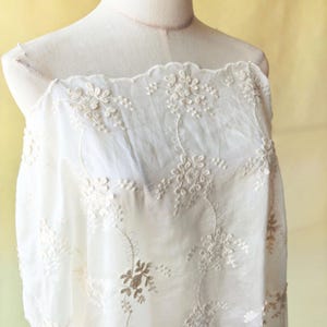 May include: Off-white floral embroidered fabric draped over a mannequin. The delicate embroidery features detailed flower and leaf patterns. The fabric has a scalloped edge and is semi-sheer, revealing the mannequin beneath. The background is a soft yellow.