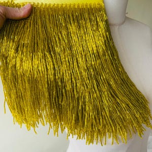 May include: Close-up of a gold beaded fringe. The fringe is made of long, thin strands of gold beads attached to a yellow fabric backing. The beads are a shiny, metallic gold color. The fringe is held by a hand.