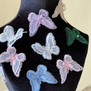 Op de afbeelding: Een verzameling delicate vlinderapplicaties in verschillende pastelkleuren, waaronder roze, blauw, groen en wit. Elke vlinder heeft ingewikkelde geborduurde details en een klein parelaccent, perfect voor knutselen of kleding.
