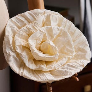 May include: A large, off-white fabric flower accessory with a crinkled, textured surface. The flower features multiple layers of gathered, pleated material arranged in a circular, blooming rose-like pattern with a soft, matte finish.