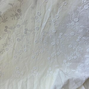 May include: Close-up of white fabric with delicate floral embroidery. The embroidery features detailed vines with small flowers and leaves, creating a textured pattern across the cloth. The fabric appears soft and slightly wrinkled.