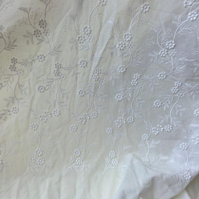 May include: Close-up of white fabric with delicate floral embroidery. The embroidery features detailed vines with small flowers and leaves, creating a textured pattern across the cloth. The fabric appears soft and slightly wrinkled.