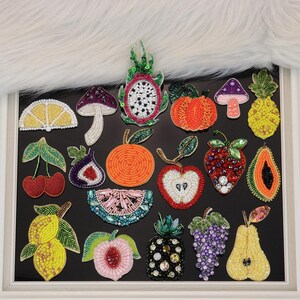 May include: A collection of twenty-one assorted fruit and mushroom shaped patches crafted with colorful beads and sequins. The set includes depictions of a dragon fruit, pineapple, strawberry, orange, lemon, pear, cherry, peach, fig, apple, and watermelon.