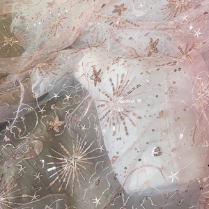 May include: Delicate, sheer fabric with a celestial design. The fabric is a soft white with rose gold embellishments, including stars, sequins, and radiating bursts. Ideal for bridal wear or special occasion garments.
