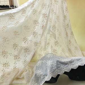 Pu&ograve; includere: Tessuto bianco sporco con un delicato motivo floreale in una tonalit&agrave; pi&ugrave; chiara. Il tessuto &egrave; drappeggiato, mostrando la sua consistenza e gli intricati dettagli dei fiori ricamati. &Egrave; visibile anche un pezzo di tessuto separato con un bordo smerlato.