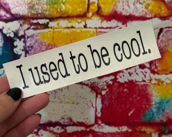 May include: A rectangular white sticker with the phrase "I used to be cool." in black, bold, sans-serif font. The sticker is held in a hand with a black painted fingernail. The background is a colorful brick wall.