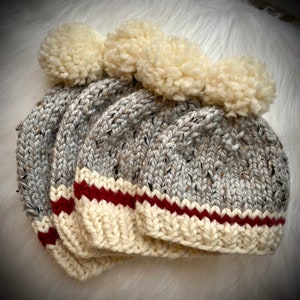 May include: Four gray and white knit beanies with cream-colored pom-poms. Each hat features a cream-colored brim with a red stripe. The hats are made of a speckled yarn.