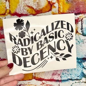 May include: A white sticker with the words "RADICALIZED BY BASIC DECENCY" in a black, stylized font. The design includes floral and star accents. The sticker is held against a colorful brick wall background.