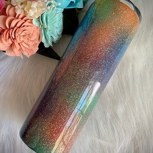 Puede incluir: Un vaso cilíndrico con un acabado de purpurina arcoíris, que se mezcla del verde al morado, naranja y amarillo. El vaso tiene una tapa transparente. Un ramo de flores de colores pastel está en el fondo.