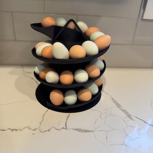 Puede incluir: Un soporte negro para huevos de tres niveles lleno de huevos marrones, blancos y azules. El soporte est&aacute; hecho de un material negro mate y se encuentra sobre una encimera blanca. Los huevos son de varios tonos.