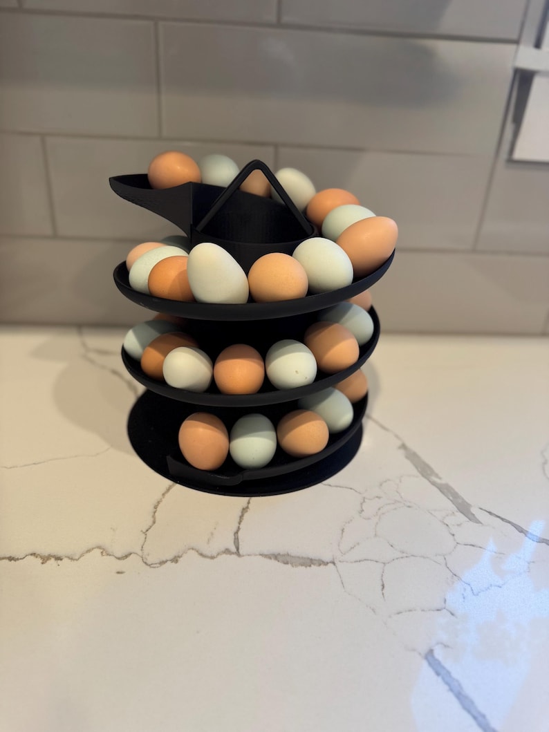 Puede incluir: Un soporte negro para huevos de tres niveles lleno de huevos marrones, blancos y azules. El soporte est&aacute; hecho de un material negro mate y se encuentra sobre una encimera blanca. Los huevos son de varios tonos.