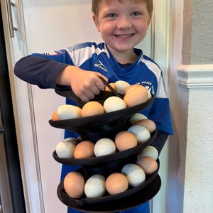 May include: A black, three-tiered egg holder filled with brown and white eggs. The egg holder is being held by a person wearing a blue soccer jersey. The eggs are arranged on circular shelves.