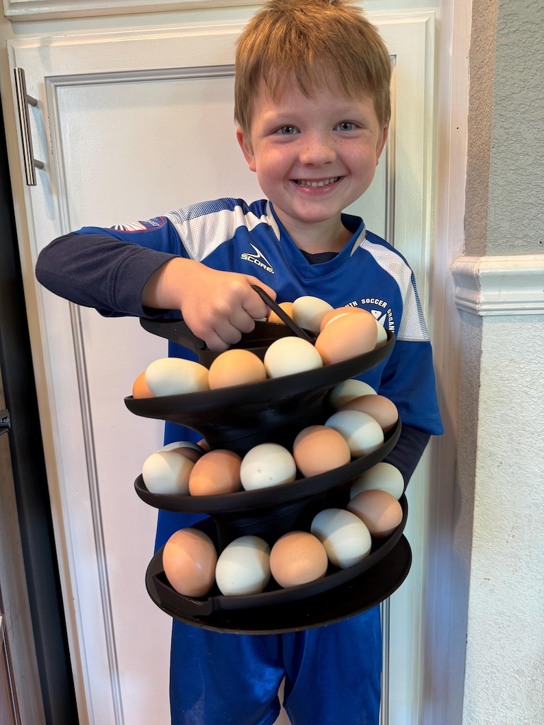 Puede incluir: Un soporte negro para huevos de tres niveles lleno de huevos marrones y blancos. El soporte para huevos lo sostiene una persona que lleva una camiseta de f&uacute;tbol azul. Los huevos est&aacute;n colocados en estantes circulares.