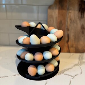 Puede incluir: Un soporte negro para huevos de tres niveles lleno de una variedad de huevos en tonos marrones, azules y blancos. El soporte es de metal y tiene un dise&ntilde;o circular. Los huevos est&aacute;n dispuestos en espiral.