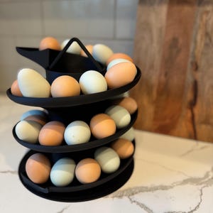 Puede incluir: Un soporte negro para huevos de tres niveles lleno de huevos en tonos marrones, azules y blancos. El soporte tiene un dise&ntilde;o moderno y se exhibe sobre una encimera blanca. Una tabla de cortar de madera est&aacute; en el fondo.