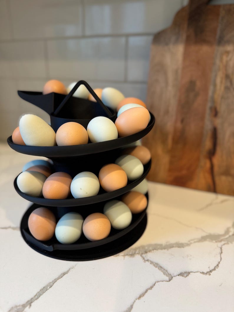 Puede incluir: Un soporte negro para huevos de tres niveles lleno de huevos en tonos marrones, azules y blancos. El soporte tiene un dise&ntilde;o moderno y se exhibe sobre una encimera blanca. Una tabla de cortar de madera est&aacute; en el fondo.