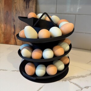 Puede incluir: Un soporte negro para huevos de tres niveles lleno de una variedad de huevos en tonos blancos, azul claro y marr&oacute;n. El soporte tiene un asa en la parte superior y est&aacute; dise&ntilde;ado para exhibir huevos. Los huevos est&aacute;n dispuestos en bandejas circulares.