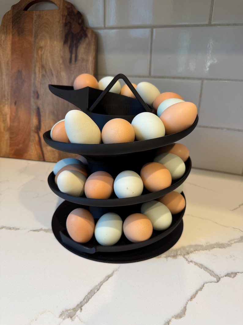Puede incluir: Un soporte negro para huevos de tres niveles lleno de una variedad de huevos en tonos blancos, azul claro y marr&oacute;n. El soporte tiene un asa en la parte superior y est&aacute; dise&ntilde;ado para exhibir huevos. Los huevos est&aacute;n dispuestos en bandejas circulares.