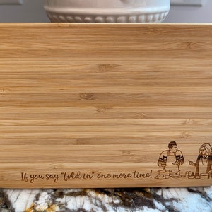 May include: Rectangular bamboo cutting board with rounded corners. The board features engraved text that reads "If you say 'fold in' one more time!" and a cartoon illustration of two people cooking.