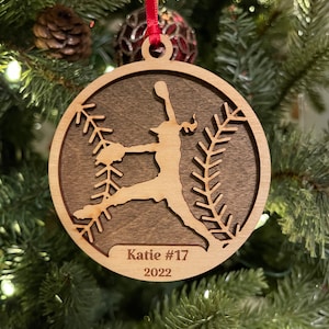 May include: A wooden Christmas ornament featuring a silhouette of a softball player in a pitching pose. The ornament is brown with a light brown silhouette and has the text "Katie #17 2022" inscribed on the bottom.