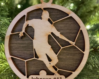 May include: A wooden soccer ornament with a silhouette of a player kicking the ball. The ornament is brown with a wood grain finish and has the text "Johnson #41 2021" on the bottom.