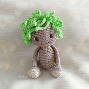Könnte beinhalten: Eine gehäkelte Puppe mit einem grünen, haarartigen Oberteil und einem grauen Körper. Die Puppe hat ein lächelndes Gesicht mit gestickten Augen und einem Blumendesign auf der Brust.