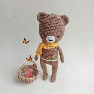 May include: A brown crocheted teddy bear wearing a yellow scarf. The bear has a white nose and embroidered eyes. The bear is standing on a white surface.