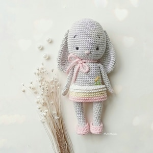 Puede incluir: Muñeca de conejo de ganchillo hecha a mano en gris suave con orejas largas y un lazo rosa. El conejo lleva una falda con rayas pastel y zapatos rosas. Una pequeña flor amarilla está bordada en el pecho. Un artículo encantador y artesanal.